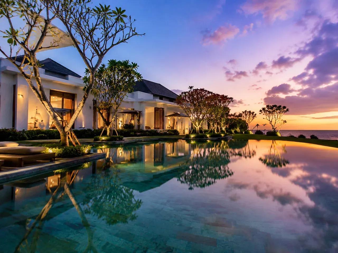 A resort infinity pool at dusk in Nusa Dua with white villas behind manicured frangipani trees
