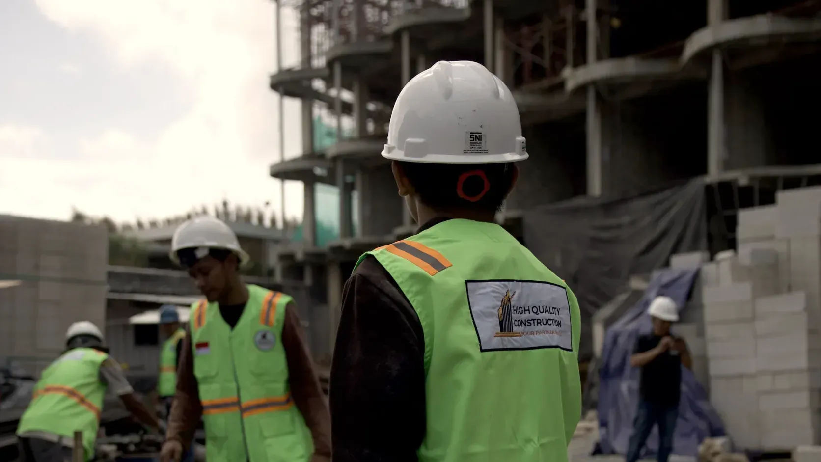 HQC site engineer in a white hard hat and High Quality Construction safety vest watching a crew at work beside a multi-storey concrete frame