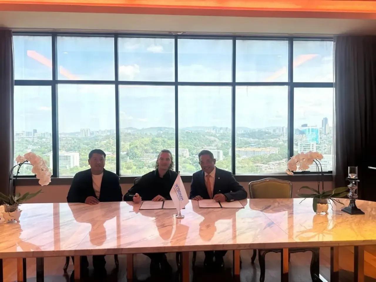 Three executives signing agreements at a long marble table in front of a panoramic window, at the signing of the Wyndham agreement for Ardhana