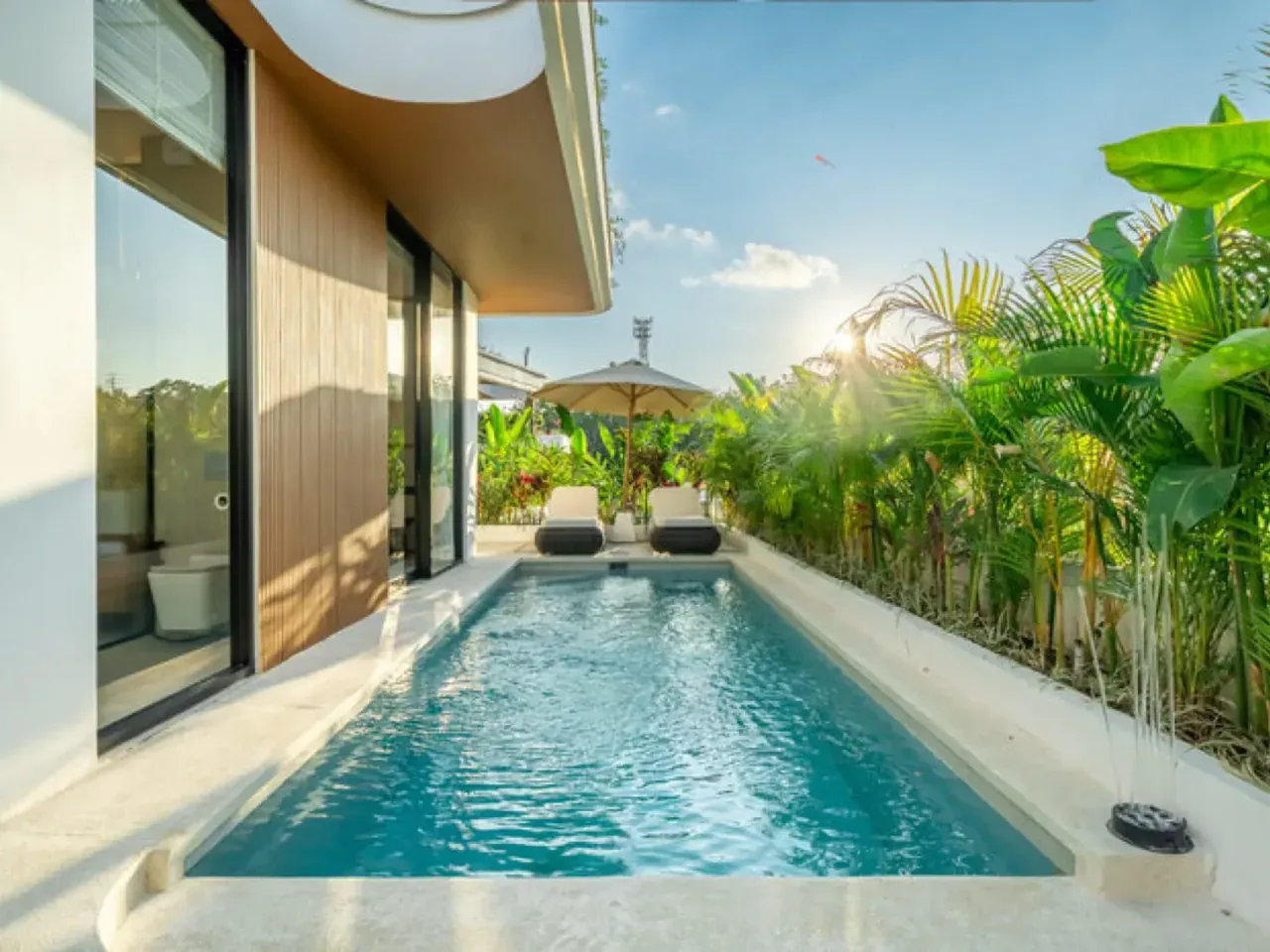 Long private swimming pool beside a villa with floor-to-ceiling glass, sun loungers and a palm garden in Bali